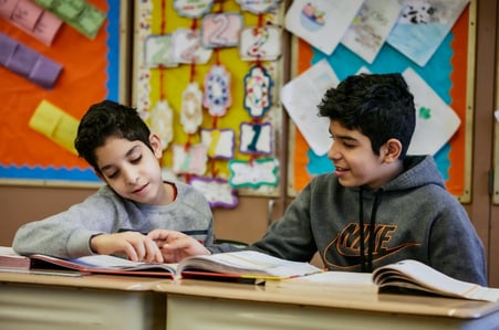 Two students point to a page of a book spread across their desks.  Two students point to a page of a book spread across their desks.